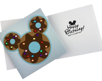 May include: A blue card with a brown and white donut shaped like Mickey Mouse's head. The card has the text "Happy Birthday! Hope it's a sweet one!" on the inside.