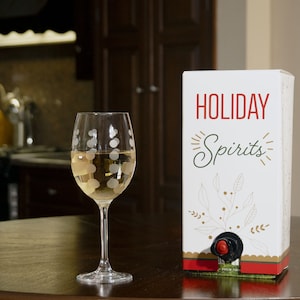 May include: A white box of wine with a red and gold design. The box says "HOLIDAY Spirits" on the front. A glass of white wine is on a wooden table next to the box.
