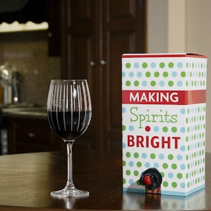May include: A box of red wine with a white and green polka dot pattern. The box says "Making Spirits Bright". A glass of red wine is on a wooden table.