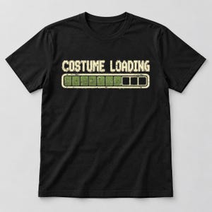 May include: A black short-sleeved t-shirt featuring the text "COSTUME LOADING" in a distressed, pixelated font. Below the text is a horizontal progress bar graphic with green filled segments, indicating partial completion.