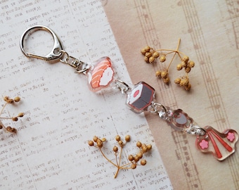 May include: A silver keychain with a charm featuring a pink and red sushi roll, a gray and white sushi roll, and a pink and orange salmon nigiri. The charm is attached to a silver key ring.