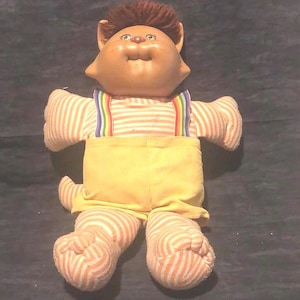 May include: A vintage plush cat doll with a tan face and brown hair. The doll wears a striped shirt with rainbow suspenders and yellow overalls. Its arms, legs, and tail are striped in orange and white. The doll has a smiling expression.