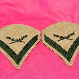 May include: Two tan and green military patches, each with a chevron design and crossed rifles. The patches are set against a bright pink background. The patches are likely for a military uniform.