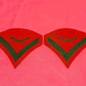 May include: Two red and green military patches. Each patch has a red background with a green chevron and crossed rifles. The patches are likely for a military uniform and are displayed on a pink background.