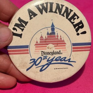 May include: A vintage Disneyland 30th Year button with the text "I'M A WINNER!". The button features a castle graphic in red and blue, with the Disneyland logo. The button is off-white with red and blue stripes.