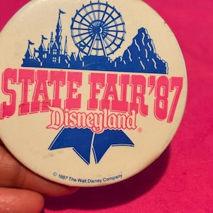 May include: A vintage button with a white background featuring a pink and blue design. The text reads "STATE FAIR '87 Disneyland" with a castle, Ferris wheel, and mountain illustration. The bottom text reads "© 1987 The Walt Disney Company."