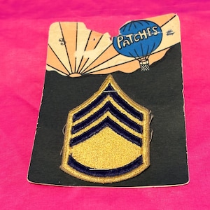 May include: A vintage gold and navy blue military patch with three chevrons, mounted on a black card with the word "PATCHES" and a hot air balloon graphic. The card has a sunburst design.