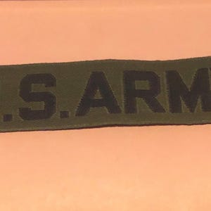 May include: Olive green rectangular patch with the text "U.S. ARMY" in black block letters. The patch is slightly worn and has a textured appearance. The background is a solid peach color.