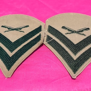May include: Two tan and green military rank insignia patches. Each patch features three dark green chevrons and a crossed rifle emblem. The patches are set against a bright pink background.