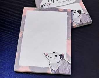 Puede incluir: Dos blocs de notas "Opossum Memo Pad". Cada bloc de notas tiene un área de escritura blanca, un borde rosa y gris y una ilustración de un mapache de dibujos animados. El bloc de notas superior muestra el texto "Opossum Memo Pad".