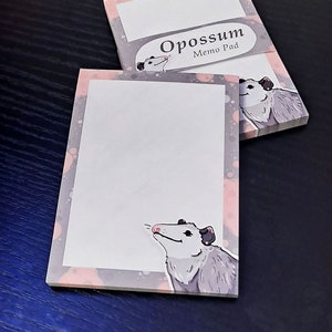 May include: Two stacks of "Opossum Memo Pad" notepads. Each notepad features a white writing area, a pink and grey border, and a cartoon opossum illustration. The top notepad displays the text "Opossum Memo Pad".