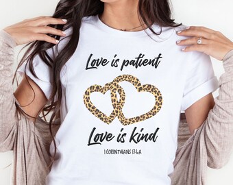 May include: A white short-sleeved t-shirt featuring two interlocking hearts with a brown and black leopard print pattern. The text reads Love is patient, Love is kind, and 1 CORINTHIANS 13:4A in a black script and sans-serif font.