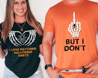 May include: Two Halloween-themed t-shirts. One black shirt features a skeleton hand heart design with the text "I LOVE MATCHING HALLOWEEN SHIRTS". The other is orange with a skeleton hand giving the middle finger and the text "BUT I DON'T".