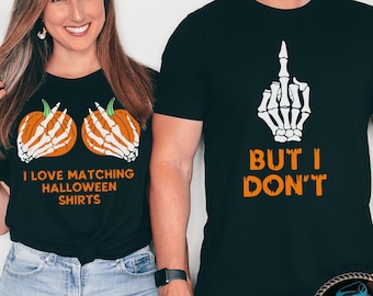 May include: A couple wearing matching Halloween shirts. The woman's shirt features two pumpkins with skeleton hands holding them and the text "I love matching Halloween shirts". The man's shirt features a skeleton hand giving the middle finger and the text "But I don't".