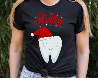 May include: Black t-shirt featuring a cartoon tooth wearing a Santa hat, with the name "Ashley" in red script above. The tooth has a smiling face. The design includes small white stars.