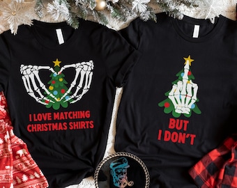 May include: Two black Christmas t-shirts. One has a skeleton hand design holding a Christmas tree with the text "I LOVE MATCHING CHRISTMAS SHIRTS". The other has a Christmas tree design with a middle finger and the text "BUT I DON'T".