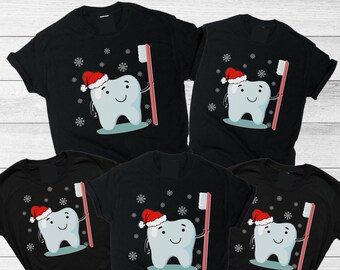 May include: Five black t-shirts featuring a graphic of a smiling white tooth character wearing a red Santa hat and holding a red toothbrush. The design includes white snowflake patterns scattered around the tooth on each shirt.