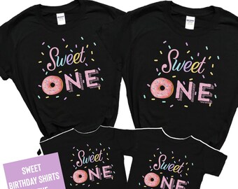 May include: Four black t-shirts featuring a graphic design with the text "Sweet ONE" in pink and white, where the letter O is replaced by a pink frosted donut with sprinkles. The design includes colorful sprinkles scattered around the text.