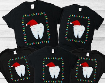 May include: Five black t-shirts with a Christmas-themed tooth graphic. Each shirt features a smiling tooth wearing a Santa hat, framed by colorful Christmas lights. The shirts are short-sleeved.