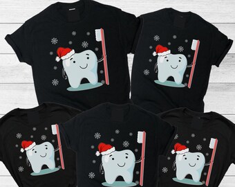 May include: Five black short-sleeved t-shirts featuring a cartoon tooth character wearing a red and white Santa hat. The tooth holds a red toothbrush, surrounded by white snowflake graphics. The design is centered on the front of each shirt.