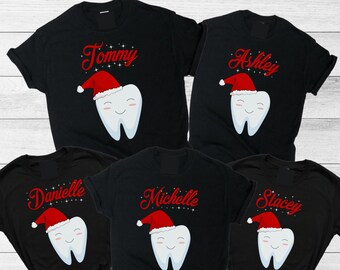 May include: Five black t-shirts with a smiling tooth wearing a Santa hat. Each shirt has a different name in red script: Tommy, Ashley, Danielle, Michelle, and Stacey. The shirts are on a light wood surface.