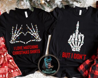 May include: Two black t-shirts featuring a skeleton hand design. One shirt reads "I love matching Christmas shirts" and the other reads "But I don't".