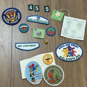 May include: A collection of Girl Scouts USA embroidered patches, pins, and insignia including a Presidential Physical Fitness Award patch, a Mid Continent strip, a Girls Are Great patch, a Camp Timberlake patch, and a Junior Girl Scout placement guide.