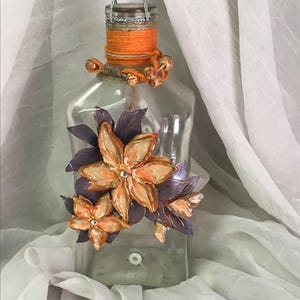 May include: A clear glass bottle with a copper-colored stopper and a decorative orange and brown twine wrap. The bottle is adorned with orange and purple floral embellishments. The stopper is attached with a silver chain.