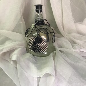 May include: A decorative glass bottle with a black stopper and wire detailing. The bottle features a black and white striped design with a black rose accent. The bottle is displayed on a white, flowing fabric.