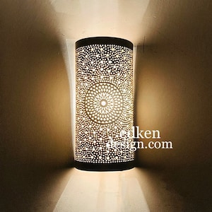 May include: A white metal wall sconce with a geometric pattern. The light is on and casting a warm glow on the wall.