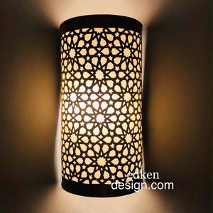 May include: A black metal wall sconce with a geometric pattern. The light is on and casting a warm glow.
