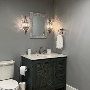 May include: A bathroom vanity with a white countertop and a dark gray cabinet with intricate carvings. The vanity has a single sink with two faucets and a mirror above it. Two decorative wall sconces with ornate metalwork and glass panels are mounted on the wall above the vanity.