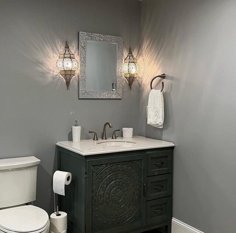 May include: A bathroom vanity with a white countertop and a dark gray cabinet with intricate carvings. The vanity has a single sink with two faucets and a mirror above it. Two decorative wall sconces with ornate metalwork and glass panels are mounted on the wall above the vanity.