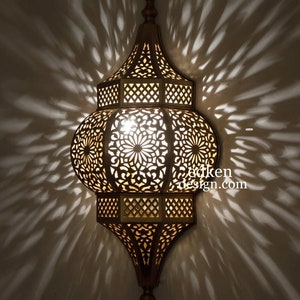 May include: A gold metal Moroccan lantern with intricate cut-out designs. The lantern is lit and casts a warm glow. The lantern is hanging from a ceiling.