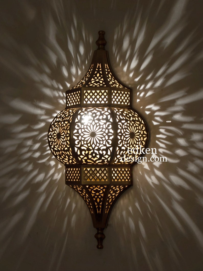 May include: A gold metal Moroccan lantern with intricate cut-out designs. The lantern is lit and casts a warm glow. The lantern is hanging from a ceiling.