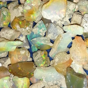 May include: A collection of rough, natural opal gemstones in various shades of white, grey, and brown. The opals have an iridescent sheen and are scattered on a dark blue surface.