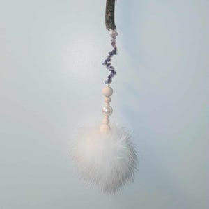 May include: A handmade cat toy featuring a natural wood stick, a string of purple beads, wooden beads, a pearl, and a fluffy white feather ball. The toy is designed to dangle and attract a cat's attention.