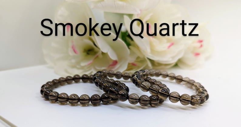 May include: Three brown smoky quartz beaded bracelets on a white surface. The bracelets are made of round beads and are stacked on top of each other. The text "Smokey Quartz" is visible above the bracelets.