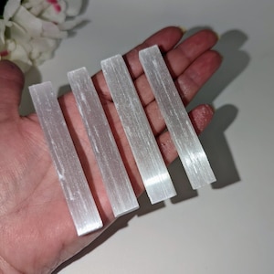 Peut inclure: Cinq bâtonnets de sélénite blanche, également appelés baguettes de sélénite, sont disposés en rangée sur une main. Les bâtonnets sont lisses et présentent une texture naturelle striée.