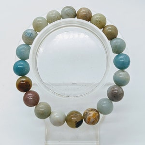 May include: A beaded bracelet featuring a variety of natural stone beads in shades of blue, brown, tan, and white. The round beads are strung together, creating a colorful and textured accessory. The bracelet is displayed on a clear stand.