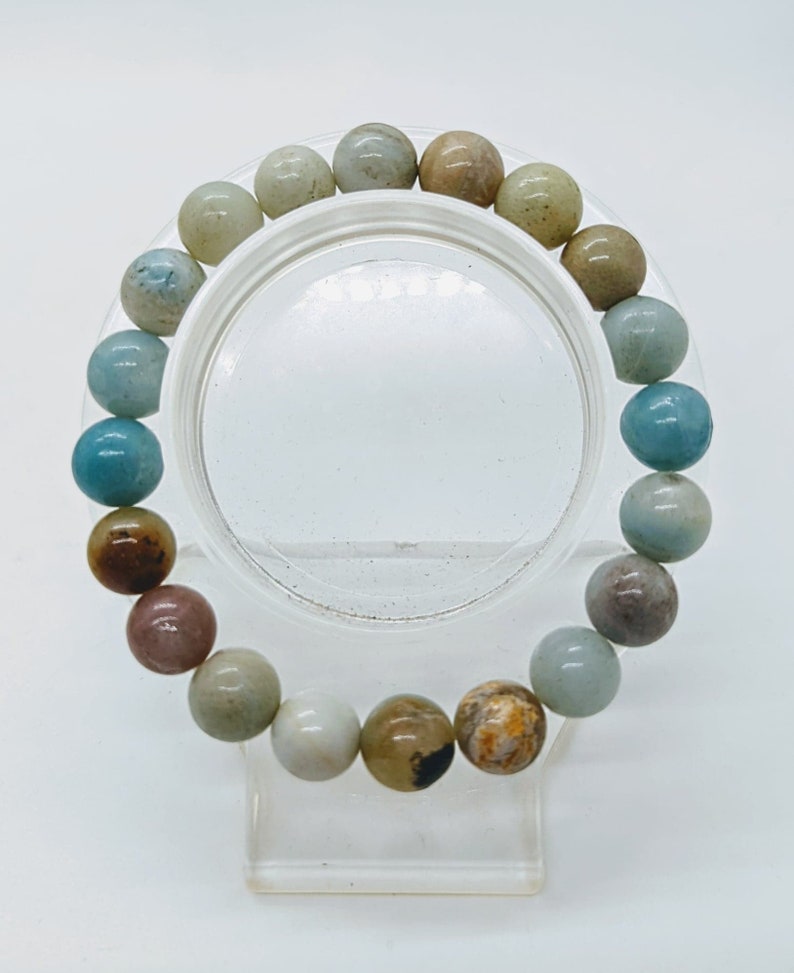 May include: A beaded bracelet featuring a variety of natural stone beads in shades of blue, brown, tan, and white. The round beads are strung together, creating a colorful and textured accessory. The bracelet is displayed on a clear stand.