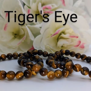 May include: Three brown tiger's eye beaded bracelets on a white surface with white flowers in the background. The text "Tiger's Eye" is visible above the bracelets.
