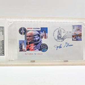 May include: A white envelope with a blue and white stamp featuring a space shuttle. The envelope is signed by John Glenn, who is also pictured on the envelope. The text "John Glenn Returns to Space" is printed on the envelope.
