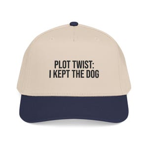 May include: A beige baseball cap with a dark blue brim and a dark blue button on top. The front of the cap features the black embroidered text PLOT TWIST: I KEPT THE DOG centered in a clean, sans-serif font across the crown of the hat.