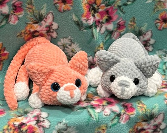 May include: Two crocheted cat toys, one in peach and one in gray, with white accents. The toys are lying on a floral patterned fabric.