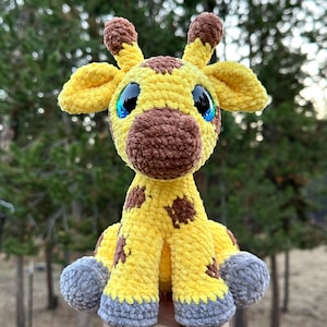 May include: A handmade, plush giraffe toy with a yellow body, brown spots, and a brown snout. The giraffe has large, blue-eyed eyes and gray feet. The toy is made of soft, textured yarn and is set against a blurred background of green trees.