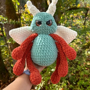 May include: A handmade crochet plush toy in the shape of a whimsical insect. The plush features a teal body, orange legs, and white wings. It has large, expressive eyes and white antennae. The toy is designed with a soft, textured yarn.