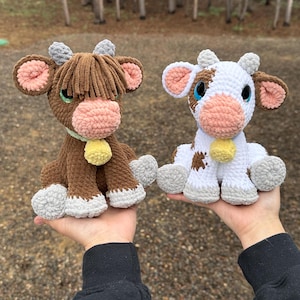 Puede incluir: Dos peluches de vaca de ganchillo. Uno es marrón con flecos, el otro es blanco con manchas marrones. Ambos tienen narices rosas, campanas amarillas y pezuñas grises. Las vacas están hechas a mano y son adecuadas para niños.