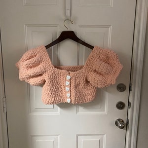 May include: A pink crocheted crop top with puffed sleeves and white heart-shaped buttons.