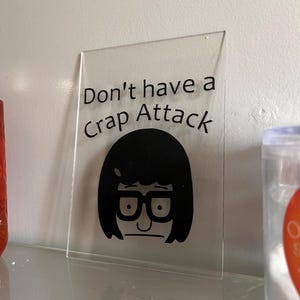 Puede incluir: Un letrero de acrílico transparente con la frase "Don't have a Crap Attack" en texto negro. Debajo del texto hay una imagen de dibujos animados en negro de un personaje con gafas y pelo corto.